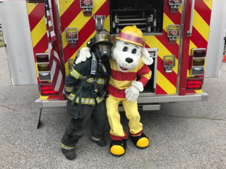 Firefighter in gear standing next to Sparky the dog behind an engine